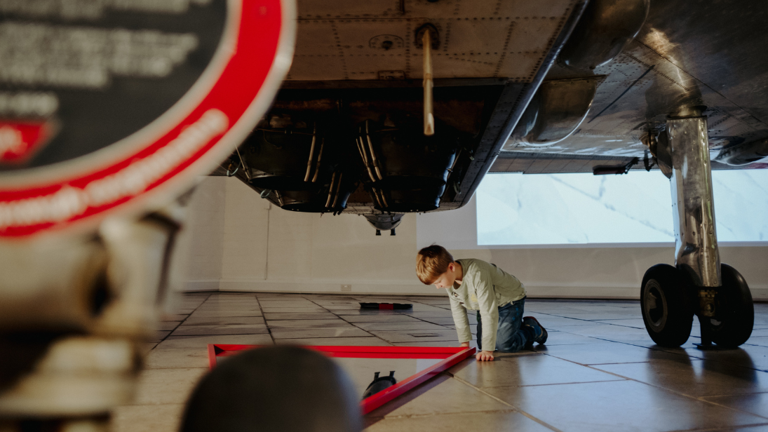 Young child on his knees on the ground, looking into a mirror placed underneath a plane exhibited in the Ulster Transport Museum.