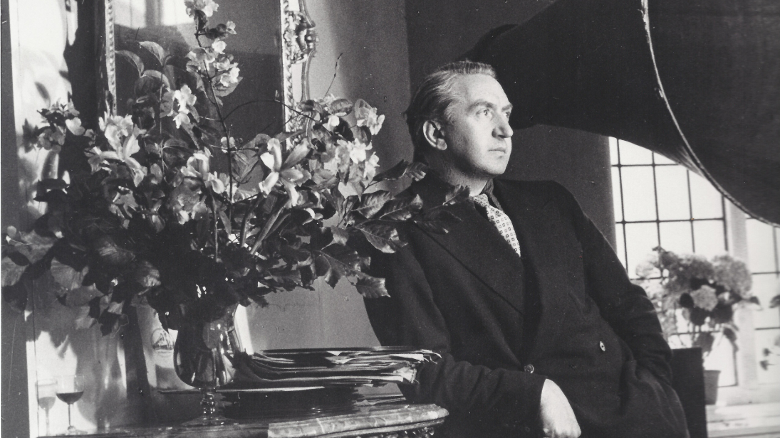 Black and white image of film director Brian Desmond Hurst, dressed in a dark suit, leaning on a side table with a vase of flowers on top, framed mirror on the wall and large gramaphone level with Hurst's head