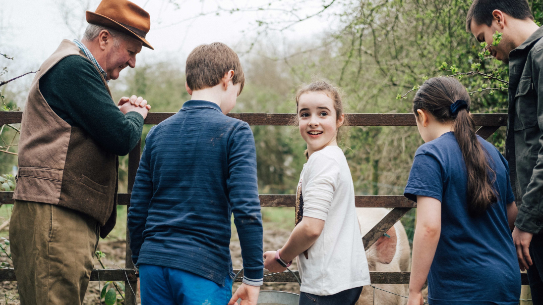 Farmer leaning on a gate with three kids standing beside him, one dressed in a long sleeve blue top with back to camera, another in a white t-shirt and this kid is look round at the camera, holding a bucket being used to feed a pig. Another child with long dark hair in a ponytail has her back to the camera, wearing a blue t-shirt. Another adult is also just in shot, wearing a coat and looking down at the pig behind the gate.