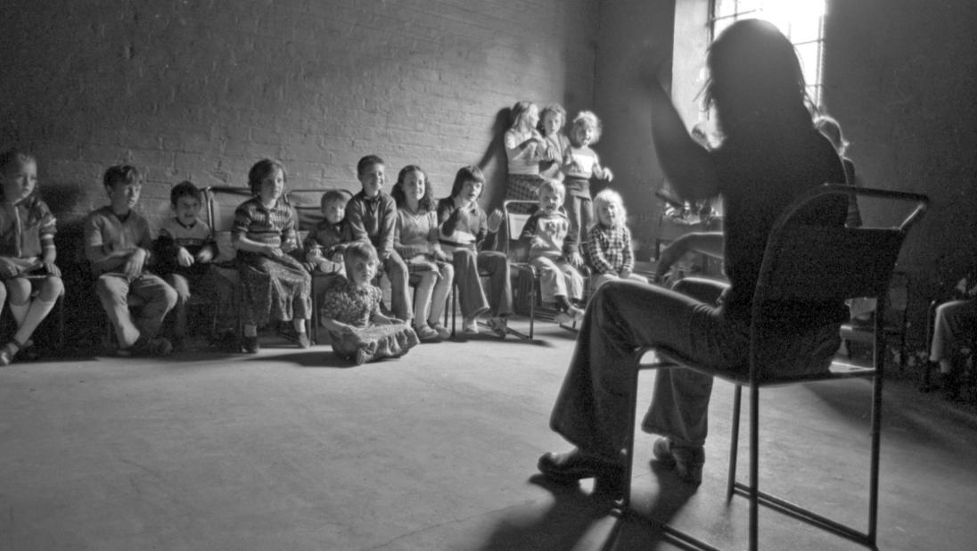 Teacher in the foreground talking and gesturing to children 