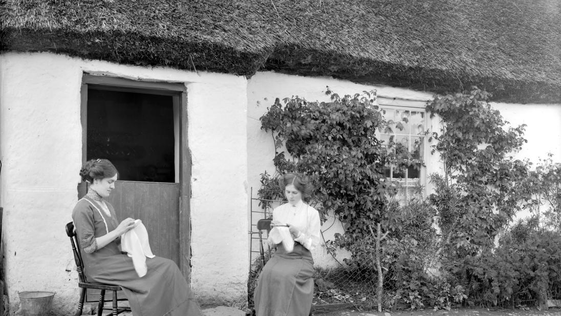 Two girls embroidering linen outside thatched house