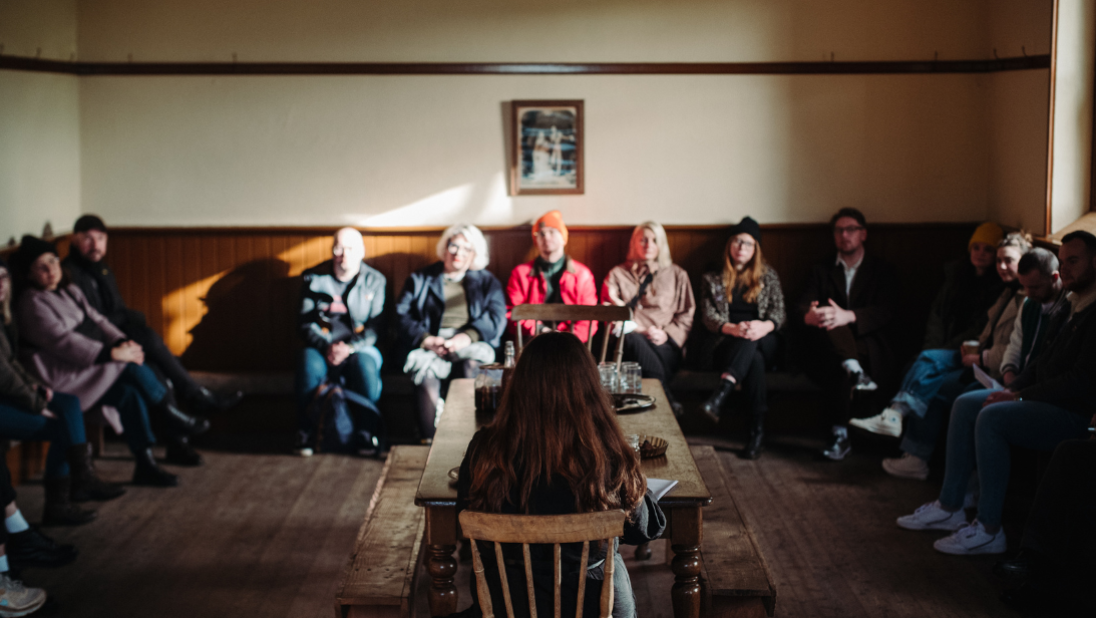 Group of people sitting on benches around a room listening to a poet