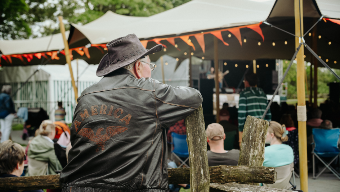 A man in a leather jacket and cowboy hat, leaning on a post listening to a performer on stage.