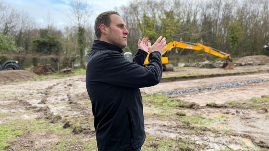 A man stands in the foreground of a field with a yellow digger in the background