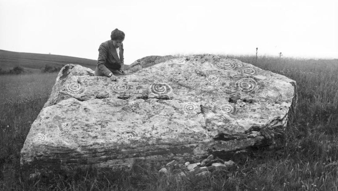 A woman sits on top of a large stone, carved with ancient symbols.