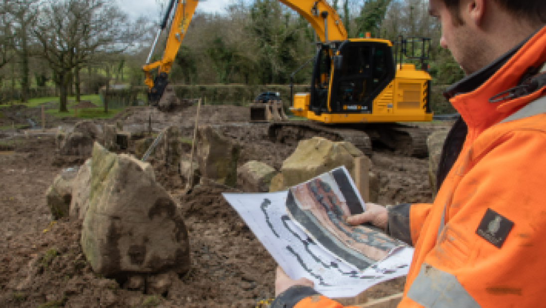 A man holds documents depicting the court tomb he is standing in front of. He wears a hard hat; in the background is a big digger.