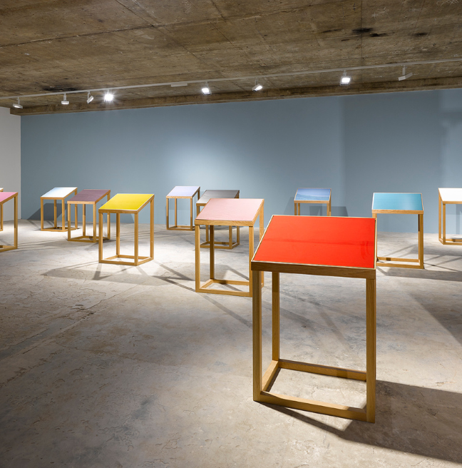 Multiple coloured topped tables - red, pink, blue, yellow tops visible - in a stone room with blue painted all at the back and lighting rack above.