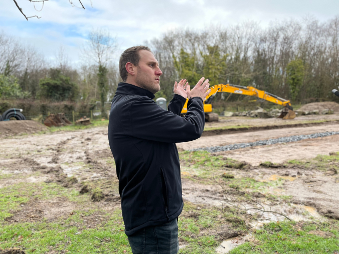 A man stands in the foreground of a field with a yellow digger in the background