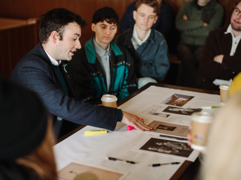 People gathered around a table with photos and paper spread on top and someone leading the conversation