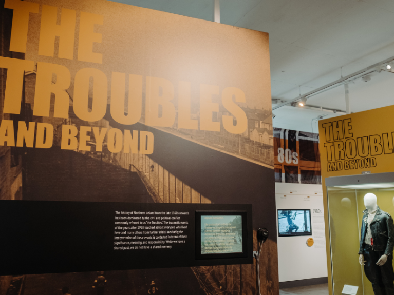 Inside the Troubles and Beyond exhibition at Ulster Museum. Yellow and black graphics on the wall say 'The Troubles and Beyond' and there is a glass display case in the background with a jacket artefact.