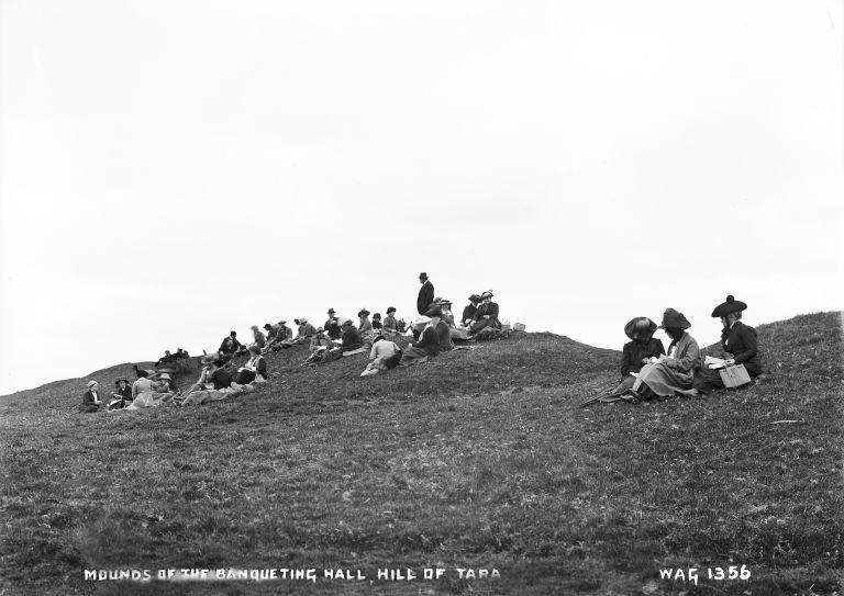 A group of people in Edwardian/Victorian dress sit for a picnic on a grassy mound.
